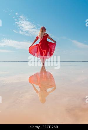Femme avec des dreadlocks colorés dans la robe rouge vif se reflète dans les eaux roses peu profondes du lac Bursol par une journée ensoleillée claire Banque D'Images