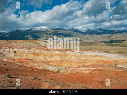 La vue aérienne capture un vaste paysage martien dans l'Altaï mettant en valeur des collines multicouches colorées et d'imposantes montagnes enneigées sous un ciel nuageux Banque D'Images