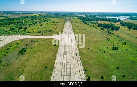 Vue aérienne d'une longue piste en béton fissuré sur un aérodrome abandonné, flanquée de champs verts et d'arbres denses. Banque D'Images