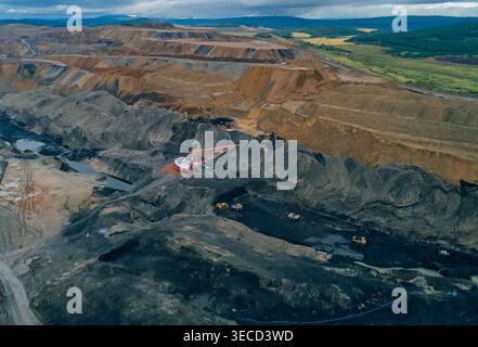 La vue panoramique aérienne capture l'exploitation minière à l'échelle industrielle avec une grande pelle hydraulique à pelle à benne traînante et plusieurs tombereaux de transport Banque D'Images