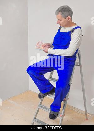 Vue verticale un plâtrier est assis sur une échelle, tenant de l'argent. Salopette bleue, chemise blanche, cheveux gris. Yeux concentrés, léger sourire, regard satisfait. Site de travail, fond propre, configuration simple. Bon pour la construction, la rénovation Banque D'Images