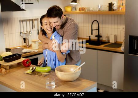 Une coréenne et un homme caucasien cuisinent dans une cuisine moderne. L'homme embrasse la femme par derrière pendant qu'elle coupe des légumes. Ils sourient et Banque D'Images