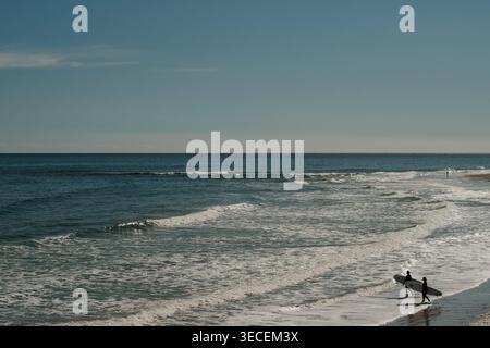 Silhouette de deux surfeurs en combinaisons avec planches de surf marchant sur la plage le long du littoral à la recherche de vagues en fin d'après-midi. Banque D'Images