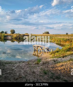 Jetée en bois rustique sur un étang calme au coucher du soleil. Une scène rurale paisible avec un quai en bois altéré s'étendant dans un étang encore réfléchissant. Banque D'Images