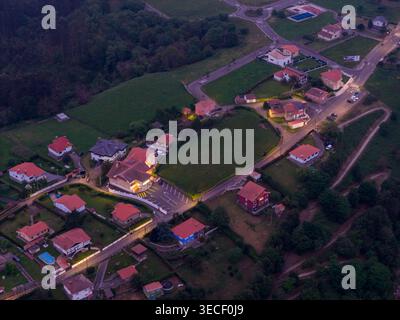 Vue aérienne de maisons illuminées aux toits rouges nichées sur une colline verdoyante au crépuscule dans le village côtier de Cudillero, Asturies, Espagne. Banque D'Images