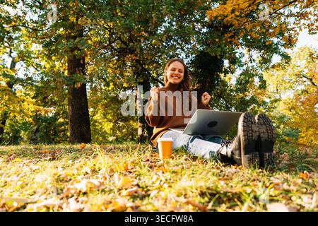 Femme joyeuse utilisant un ordinateur portable tout en étant assis sur le sol dans le parc sur le parc d'automne ensoleillé Banque D'Images
