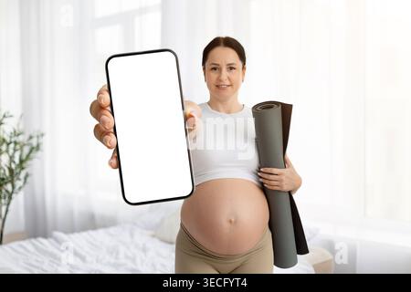Femme enceinte tenant un tapis de yoga sous le bras et montrant un écran de smartphone vierge Banque D'Images