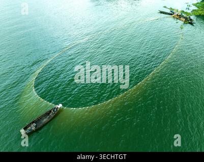 Vue aérienne de pêcheurs dans des bateaux en bois jetant un grand filet de pêche circulaire sur les eaux vertes du lac Kaptai Rangamati, division de Chittagong, Bangl Banque D'Images