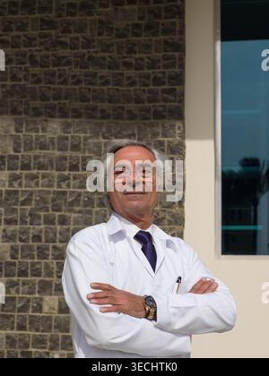 Médecin masculin senior en manteau blanc debout en toute confiance à l'extérieur de la clinique avec les bras croisés Banque D'Images