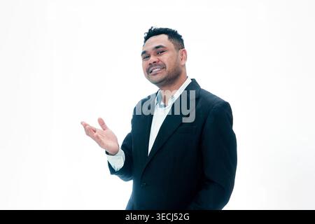 Un homme indonésien souriant en costume noir montrant un geste d'accueil ou présentant un produit, isolé sur un fond blanc propre. Banque D'Images