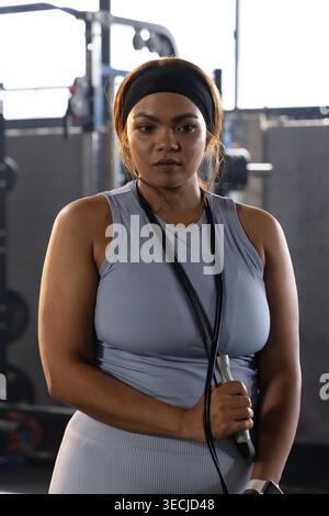 Femme afro-américaine debout tenant une corde à sauter portant une tenue bleue et un bandeau noir au gymnase Banque D'Images