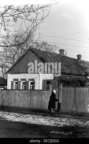Slaviansk, milieu des années 1950 - début des années 1960 Une scène paisible de quartier hivernale avec une femme âgée dans un manteau sombre et un foulard blanc ouvrant doucement la lourde porte en bois d'une clôture résidentielle. Derrière la clôture se dresse une maison classique à un étage caractérisée par son toit sombre texturé et sa bordure de fenêtre ornementale. La rue calme est légèrement recouverte de neige, reflétant le rythme calme de la vie quotidienne de banlieue. Un aperçu vraiment poignant de la vie avant la guerre, mettant en valeur la région paisible du Donbass. Banque D'Images