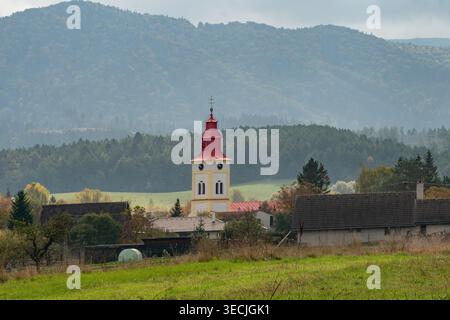Vue aérienne de la tour historique de l'église avec son toit rouge distinctif entouré de maisons traditionnelles et de montagnes boisées à Mosovce, Zilina Regi Banque D'Images