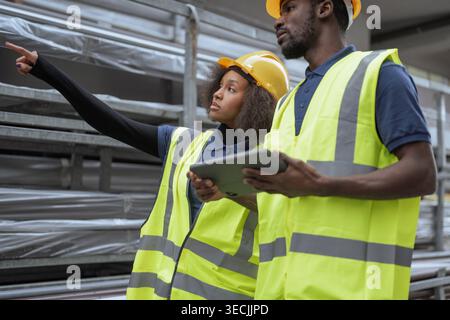 Deux travailleurs portant des vêtements de protection, des casques de sécurité jaunes et des gilets de sécurité faisant l'inventaire dans un entrepôt extérieur à l'aide d'une tablette numérique. Banque D'Images