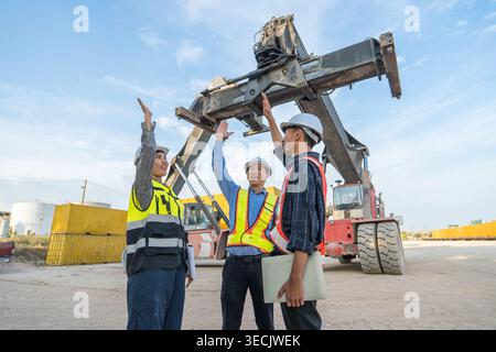 Trois professionnels de la logistique asiatique mi-adulte diversifiés en gilets de sécurité célèbrent le chargement réussi de conteneurs dans un terminal portuaire thaïlandais pour l'international Banque D'Images