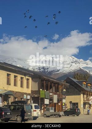 Mestia, Géorgie - 02 novembre 2025 : Mestia, Géorgie. Vue ensoleillée sur la ville. Des sommets enneigés s'élèvent au-dessus d'une rue rustique de village. Les oiseaux glissent à travers le ciel bleu vif au-dessus des toits. Les véhicules et les gens se déplacent tranquillement parmi les bâtiments en pierre Banque D'Images