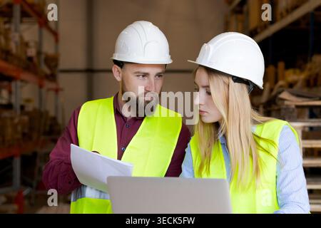 Travailleurs masculins et féminins concentrés analysant le document et l'écran d'ordinateur portable. Résolution de problèmes et pensée critique dans les opérations de gestion d'entrepôt Banque D'Images