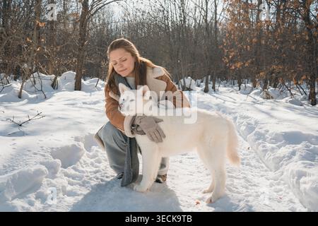 Une jeune femme embrasse chaleureusement un adorable chiot blanc Akita dans un paysage hivernal serein. Entourée d'arbres enneigés, la scène évoque le confort A. Banque D'Images