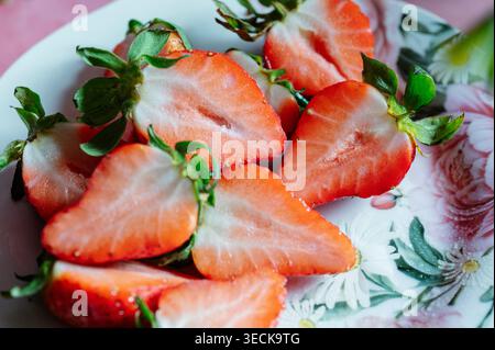 Gros plan de moitiés de fraises fraîches tranchées avec des feuilles vertes sur une assiette florale, une lumière naturelle vive et une mise au point douce, célébrant les fruits du printemps, Freshne Banque D'Images
