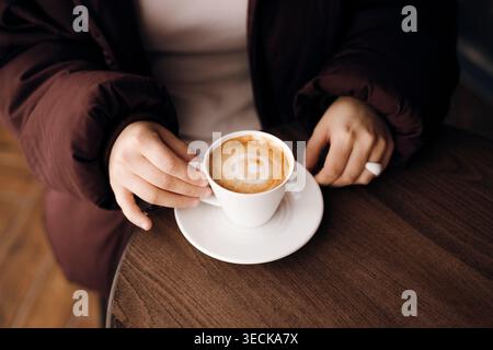 Gros plan d'une personne dans une veste chaude tenant une tasse blanche de cappuccino avec de la mousse de latte sur une soucoupe, reposant sur une table en bois dans un cadre de café confortable. Banque D'Images