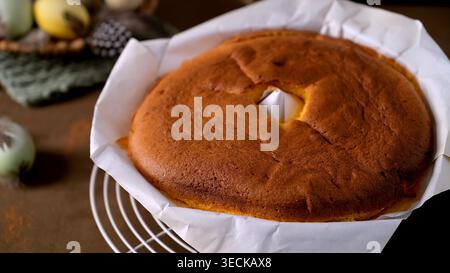 Un gâteau éponge moelleux et doré repose sur du papier parchemin au sommet d'une grille blanche. Un éclairage latéral doux met en valeur la texture poreuse contre un dos rustique Banque D'Images