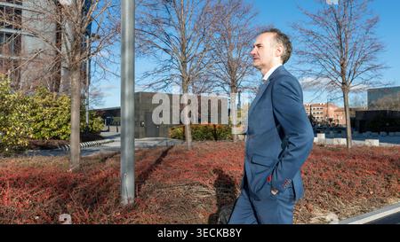 Un homme d'affaires d'âge moyen en costume bleu se promène soigneusement dans un parc urbain, les mains dans les poches, entouré d'immeubles de bureaux et de trés sans feuilles Banque D'Images