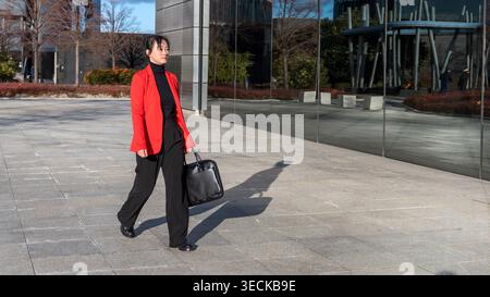 Femme d'affaires d'âge moyen marche en toute confiance à travers un paysage urbain, portant un blazer rouge et tenant une mallette noire, reflétant le professionnalisme et le pu Banque D'Images