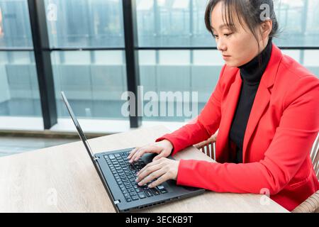 Une femme d'affaires concentrée d'âge moyen dans un blazer rouge travaille efficacement sur son ordinateur portable dans un bureau moderne avec des fenêtres en verre, symbolisant la productivité et Banque D'Images