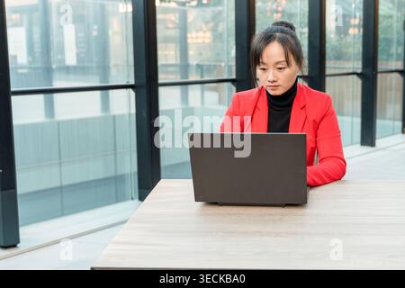 Professionnel d'affaires d'âge moyen dans un blazer rouge travaillant sur un ordinateur portable à un bureau moderne, concentré et dédié, entouré de grandes fenêtres et Banque D'Images