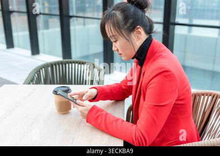 Une femme d'affaires d'âge moyen dans un blazer rouge vif est assise à une table dans un bureau moderne, engagée avec son smartphone alors qu'elle se concentre sur le travail, symbolisant Banque D'Images