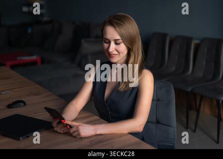 Jeune femme d'affaires assise à un bureau moderne, souriante tout en utilisant un smartphone à côté d'un ordinateur portable. Atmosphère professionnelle de l'espace de travail, productivité, Banque D'Images