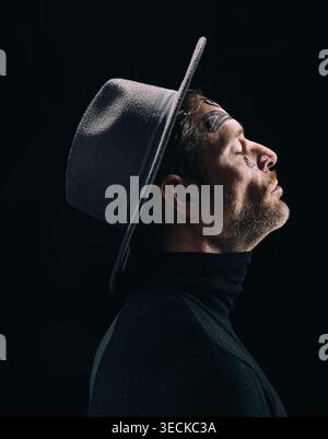 Portrait studio low key d'un homme avec tatouage facial en profil latéral, portant un chapeau à large bord et col roulé noir, regardant vers le haut contre un dos sombre Banque D'Images