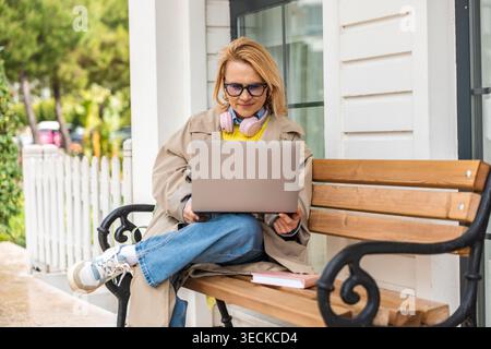 Une femme est assise à l'extérieur sur un banc en bois, embrassant un style de vie détendu. Portant des écouteurs autour du cou, elle travaille sur un ordinateur portable, profitant de la tranqu Banque D'Images