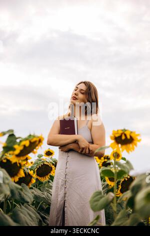 Jeune femme paisible dans un champ de tournesol tenant une Sainte Bible à sa poitrine, les yeux fermés dans la prière et la réflexion, sous la lumière douce du soir et nuageux Banque D'Images