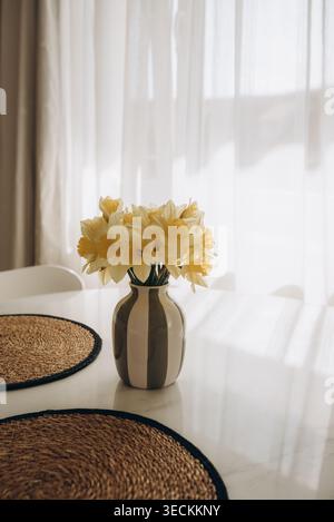 Scène intérieure minimaliste avec un bouquet de jonquilles jaunes fraîches dans un vase en céramique placé sur une table blanche. Banque D'Images
