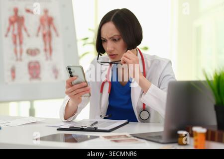 Médecin dans une clinique moderne vérifie son téléphone avec un regard inquiet tout en étant entouré de dossiers médicaux et d'un ordinateur portable en plein jour Banque D'Images