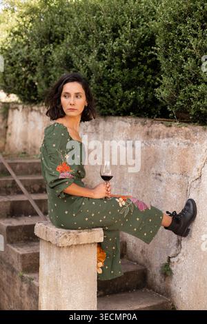 Une jeune femme aux cheveux courts est assise sur une balustrade d'un vieil escalier. Elle tient un verre de vin rouge dans une main. Le soleil brille sur elle comme elle a l'air awa Banque D'Images