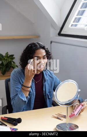 Personne non binaire appliquant le maquillage au bureau par puits de lumière à l'aide de miroir, pinceaux, palette en denim Banque D'Images