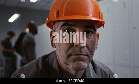 Gros plan d'un travailleur d'usine chevronné en veste de travail grise et casque orange, assis sur le banc et regardant dans le cadre avec des casiers en métal et des collègues en arrière-plan. Préparation industrielle. Portrait. Banque D'Images