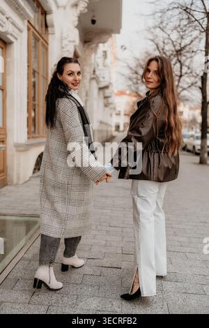 Femmes amitié marchant deux jeunes dames élégantes se tiennent la main sur une rue de la ville pendant l'heure d'or portant des vêtements d'extérieur à la mode Banque D'Images