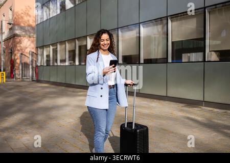 Femme d'affaires souriante vérifie smartphone tout en tirant une valise roulante le long d'un immeuble de bureaux moderne, capturant la mobilité d'affaires, les déplacements et le bus Banque D'Images