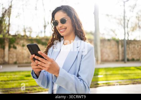 Femme d'affaires souriante dans un blazer bleu clair vérifie son smartphone à l'extérieur, portant des lunettes de soleil à la lumière du jour. Style de vie professionnel, communication mobile Banque D'Images