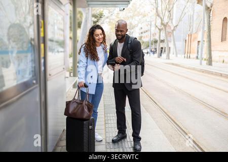 Couple d'affaires en voyage d'affaires en ville se tient à un arrêt de tram, vérifier les directions sur un smartphone. Ils voyagent avec des bagages, des sacs à dos et des bagages à main, Banque D'Images