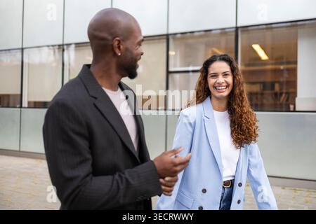 Couple d'affaires souriant dans des tenues décontractées intelligentes marchant et parlant à l'extérieur d'un immeuble de bureaux en verre moderne, capturant le travail d'équipe, le partenariat et le posit Banque D'Images
