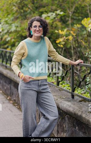 Femme mature avec des cheveux bouclés et des lunettes, vêtue de façon décontractée, profite d'un moment de détente appuyé sur une balustrade de fer dans un cadre verdoyant de parc urbain, EM Banque D'Images
