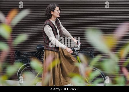 Femme mature avec les cheveux bouclés et les lunettes marche le long de sa bicyclette dans un cadre urbain. Elle est habillée dans des tons automnaux et un gilet à carreaux, appréciant Banque D'Images