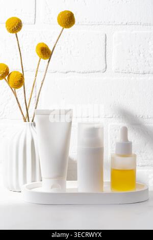 Cosmétiques de soins de la peau blancs minimaux sur un plateau rond. Nettoyez le tube, le flacon pompe et le sérum compte-gouttes en verre à côté d'un vase en céramique avec des fleurs séchées jaunes sur un Banque D'Images