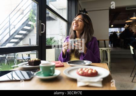 Femme souriante est assise à une table de café par une grande fenêtre, tenant une tasse de café chaud pendant un temps de café détendu, avec des pâtisseries, tablette, et dayli doux Banque D'Images