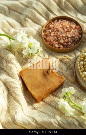 Savonnette naturelle rustique à côté d'un bol de sel de spa rose et de délicates fleurs blanches sur un tissu beige texturé, créant un bien-être chaleureux et apaisant et de soi Banque D'Images