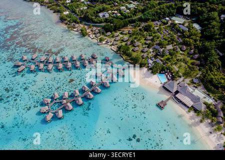 Vue aérienne d'une station balnéaire tropicale avec bungalows sur l'eau, eau turquoise, récif de corail et plage de sable blanc bordée de palmiers luxuriants en P français Banque D'Images